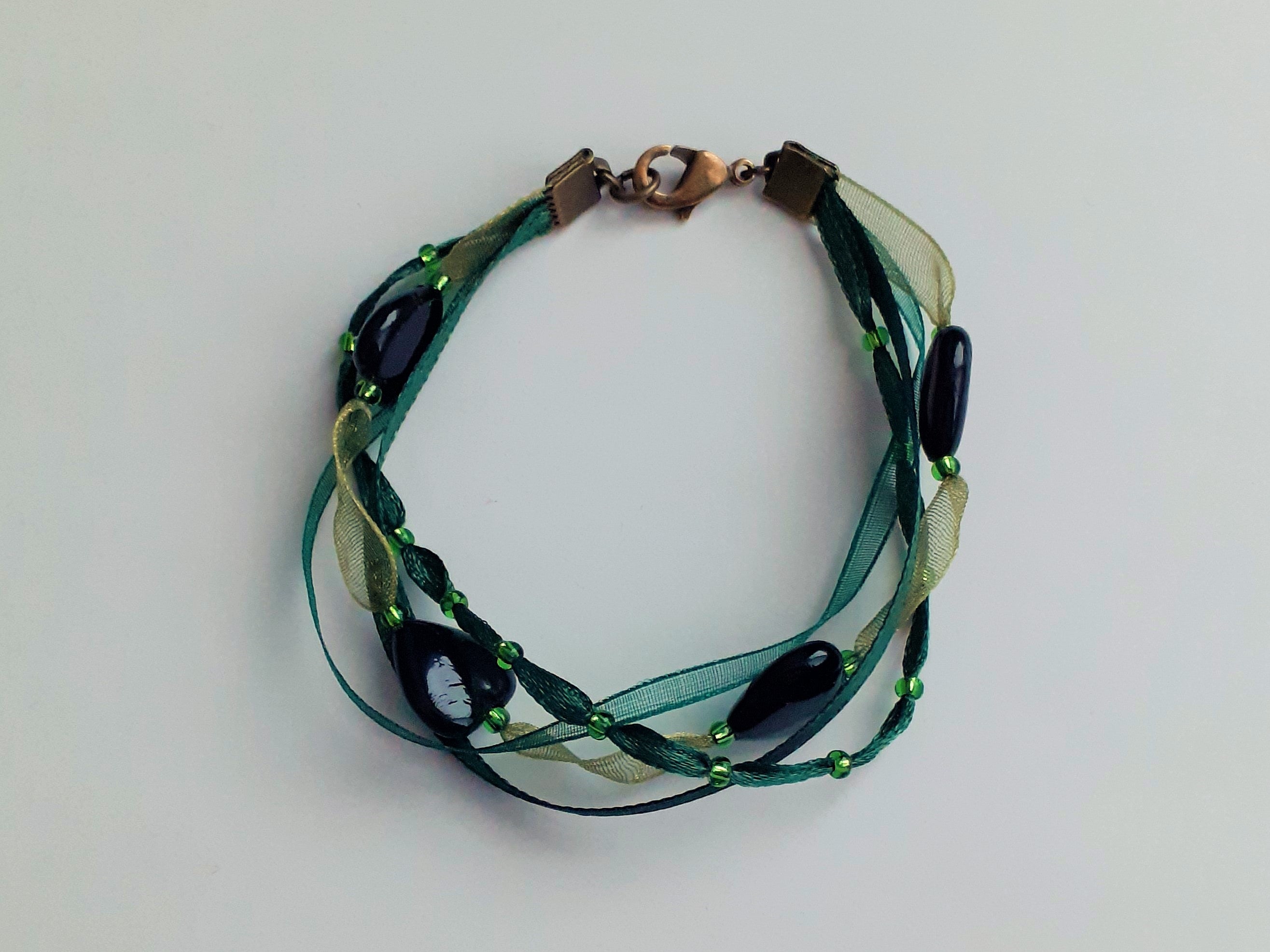 Bracelet Syléra : Trois rubans aux nuances de vert entrelacés, ponctués de perles et de cœurs profonds, pour une élégance naturelle et lumineuse au poignet.