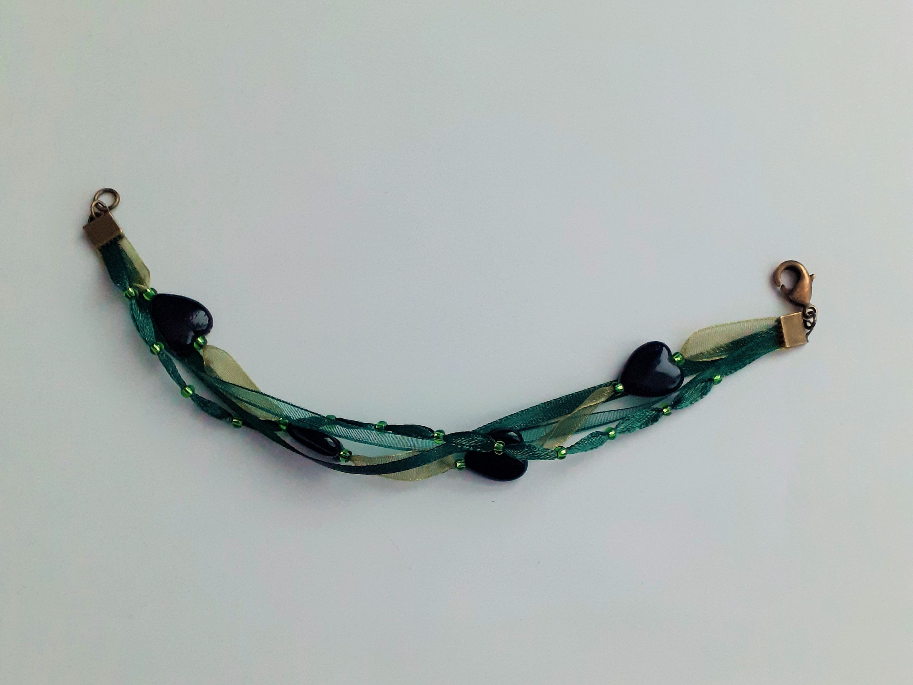 Bracelet Syléra : Trois rubans aux nuances de vert entrelacés, ponctués de perles et de cœurs profonds, pour une élégance naturelle et lumineuse au poignet.